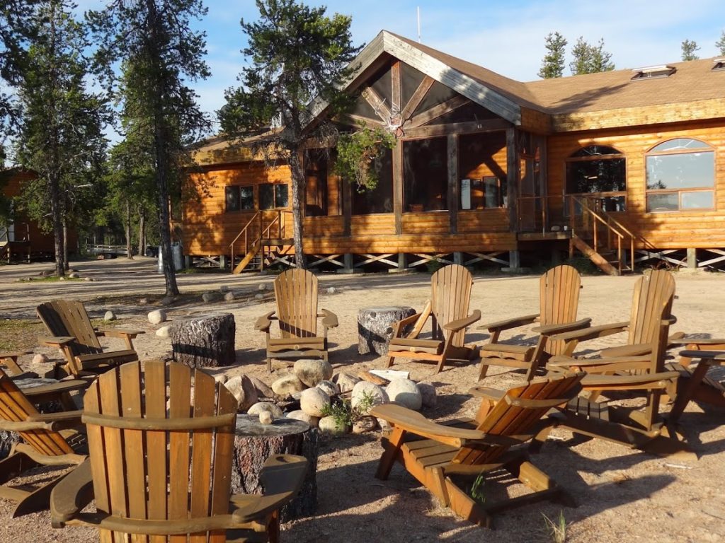 landscape image of a campfire with lounging chairs and a main wooden lodge in the background