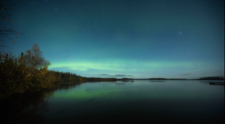 view of the northern lights over the lake and trees in the distance